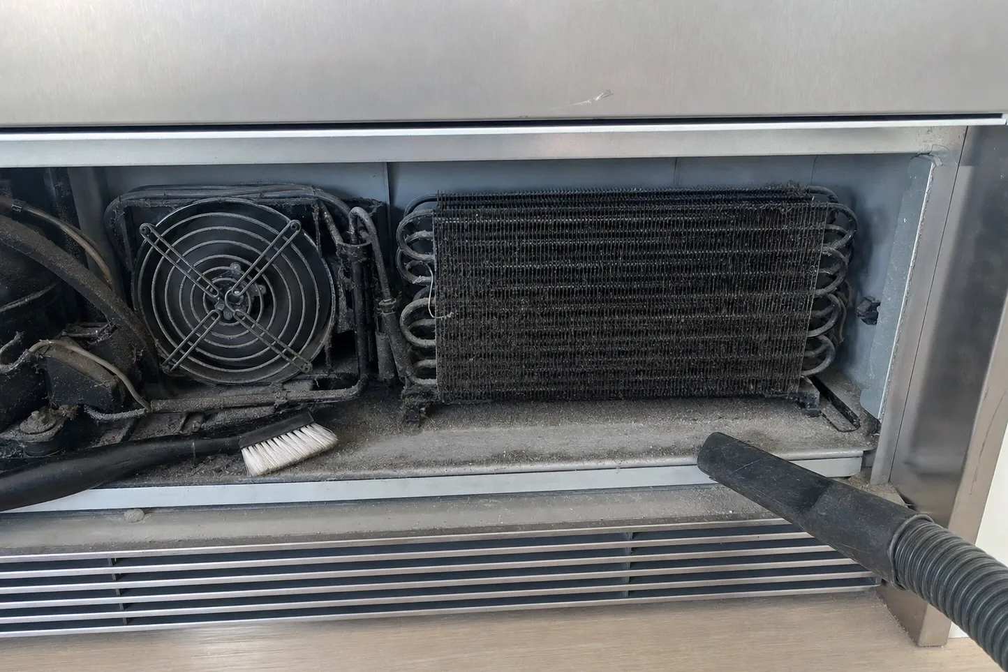 Toe-kick grille removed from a stainless built-in, showing a condenser coil and caged fan furred with grey lint, a brush inside and a vacuum nozzle at the edge