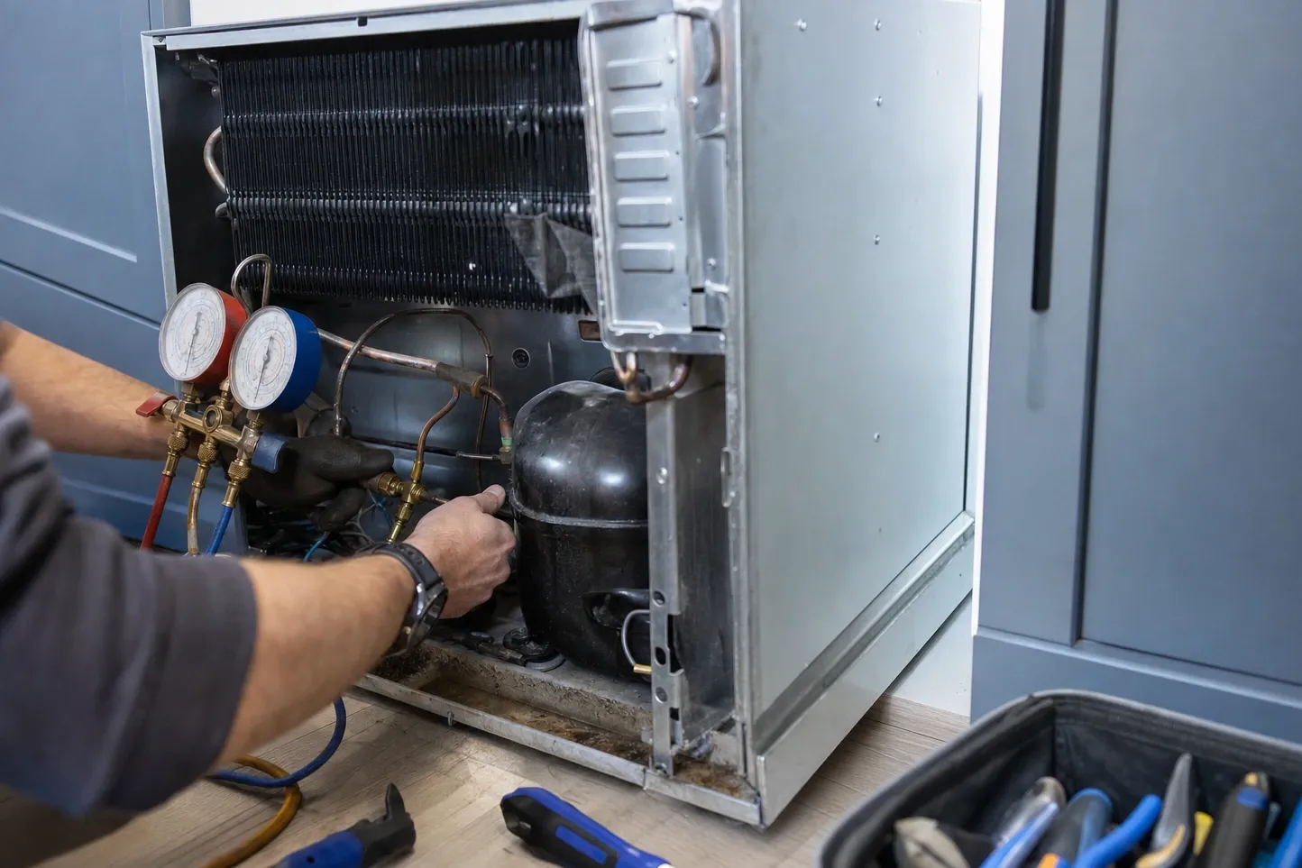 Undercounter unit pulled clear of a cabinet run with its rear panel off, condenser coil above the compressor and a manifold gauge set alongside
