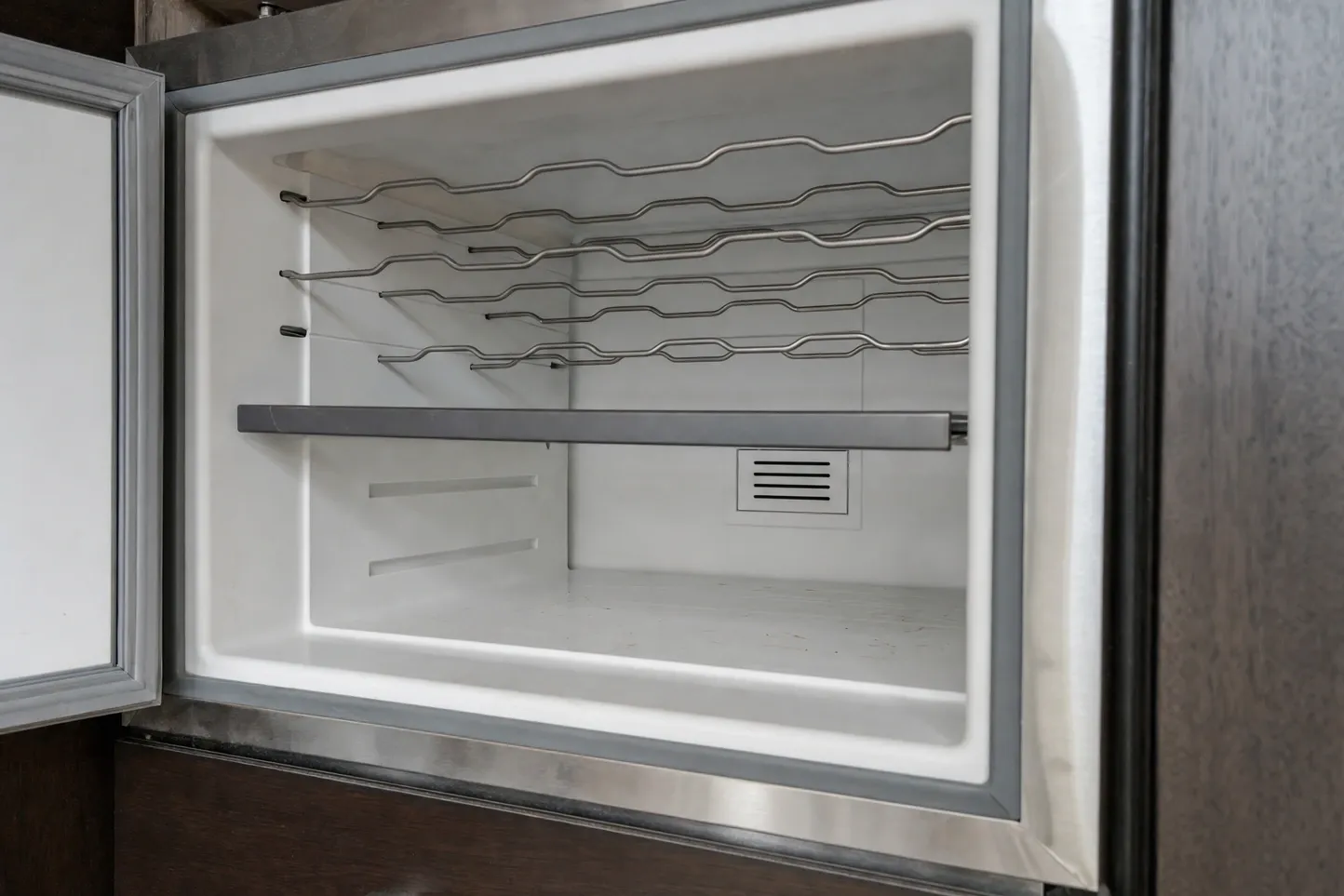 Empty undercounter interior with a white liner, three wavy wire racks above a shelf rail and a small louvred vent panel on the back wall